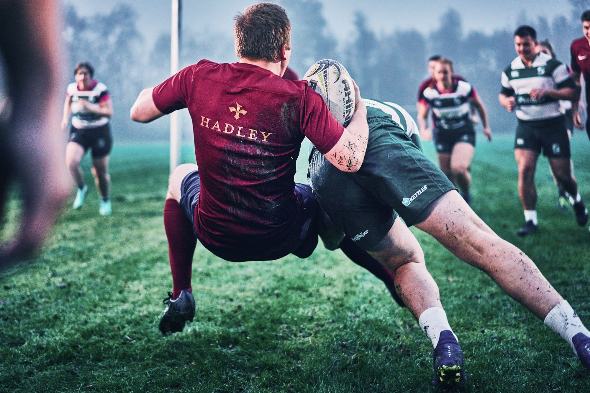 A HADLEY team player in a rugby match is tackled while teammates and opponents rush in the background.